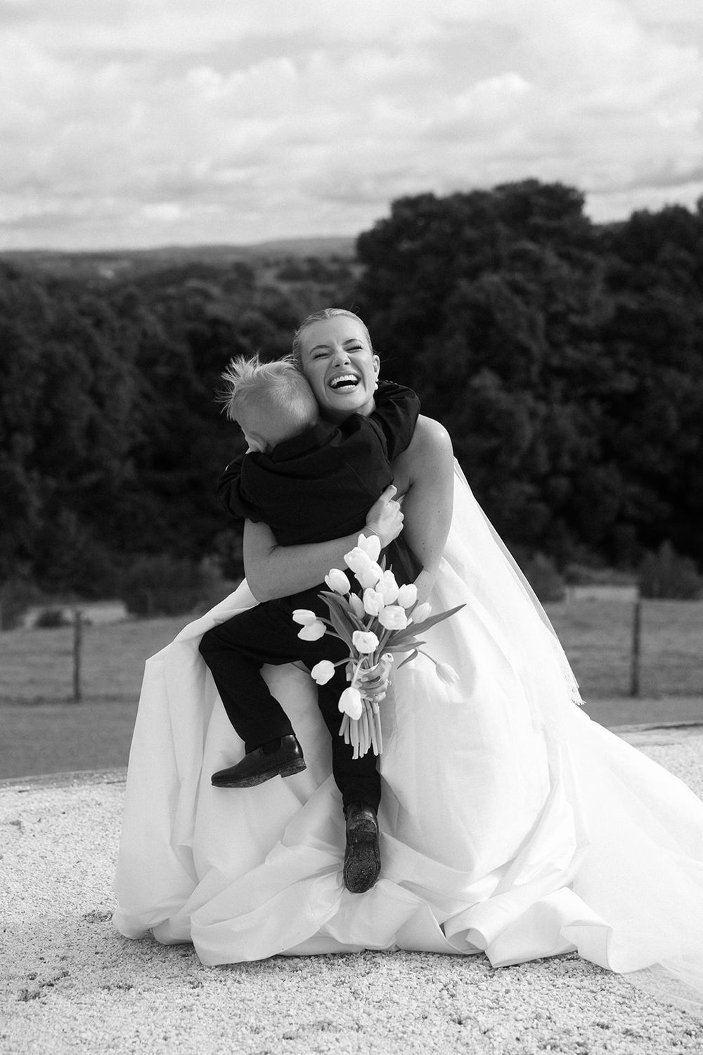 Laughing bride in white gown hugs young boy in black suit outdoors in joyful black and white wedding photo.