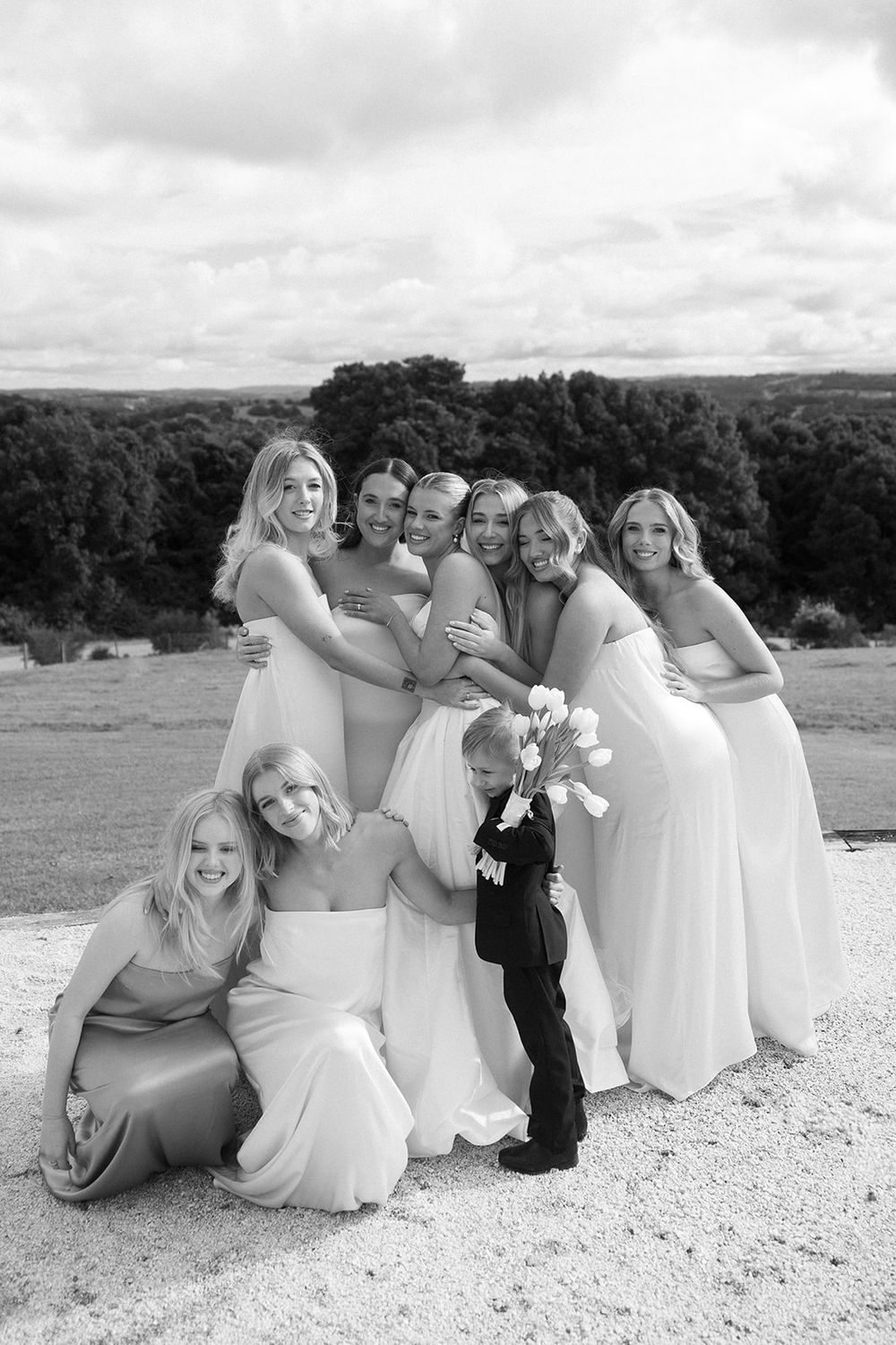 Black and white photo of bridesmaids in elegant gowns posing with a young ring bearer outdoors at a wedding.