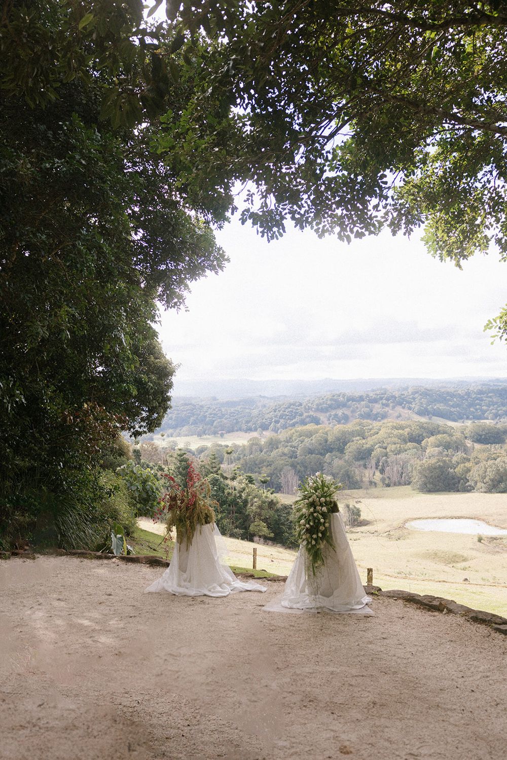 Two floral arrangements on white stands overlook a lush green valley landscape at an outdoor wedding ceremony.