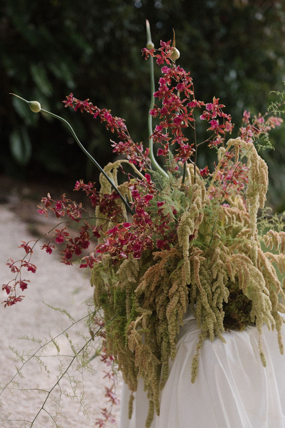 Elegant floral arrangement with cascading amaranthus and red orchids in a white vase against green foliage.