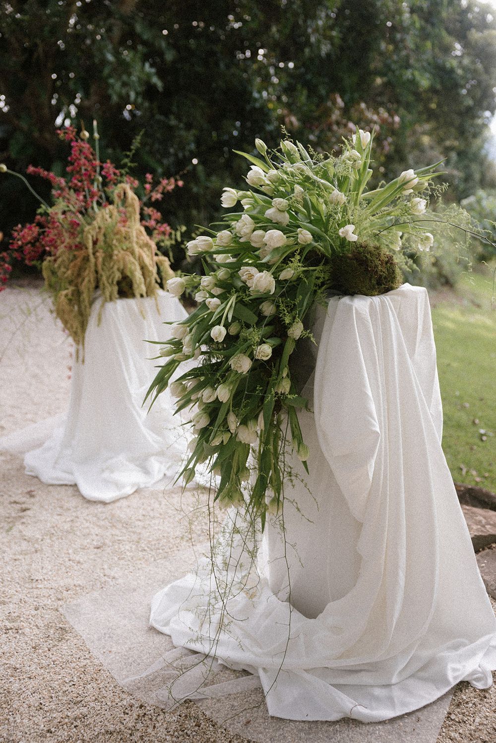 White draped pedestals adorned with lush green foliage and white tulip arrangements at an elegant outdoor wedding ceremony.