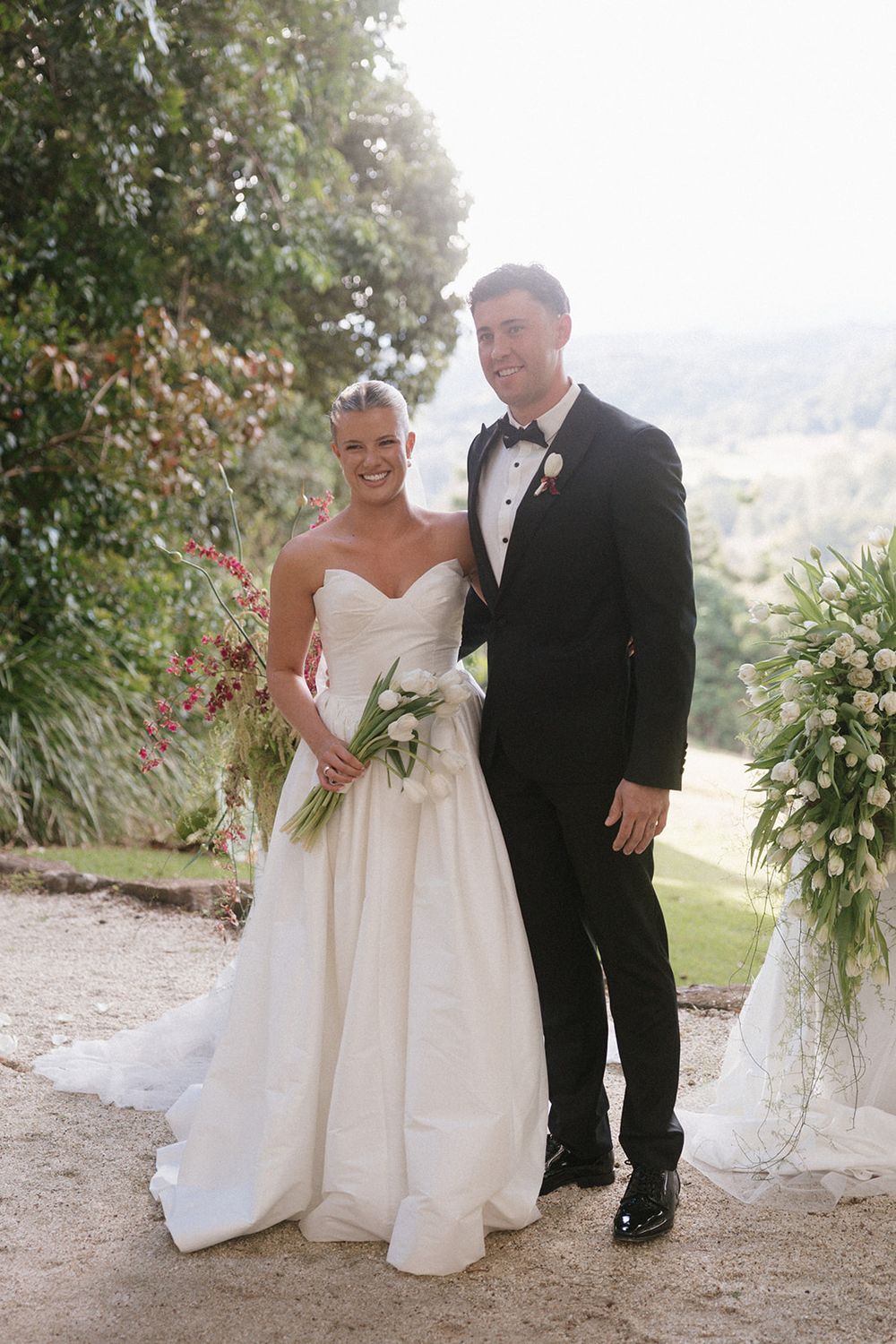 Bride in strapless white gown holding tulip bouquet stands with groom in black tuxedo outdoors at elegant wedding venue.