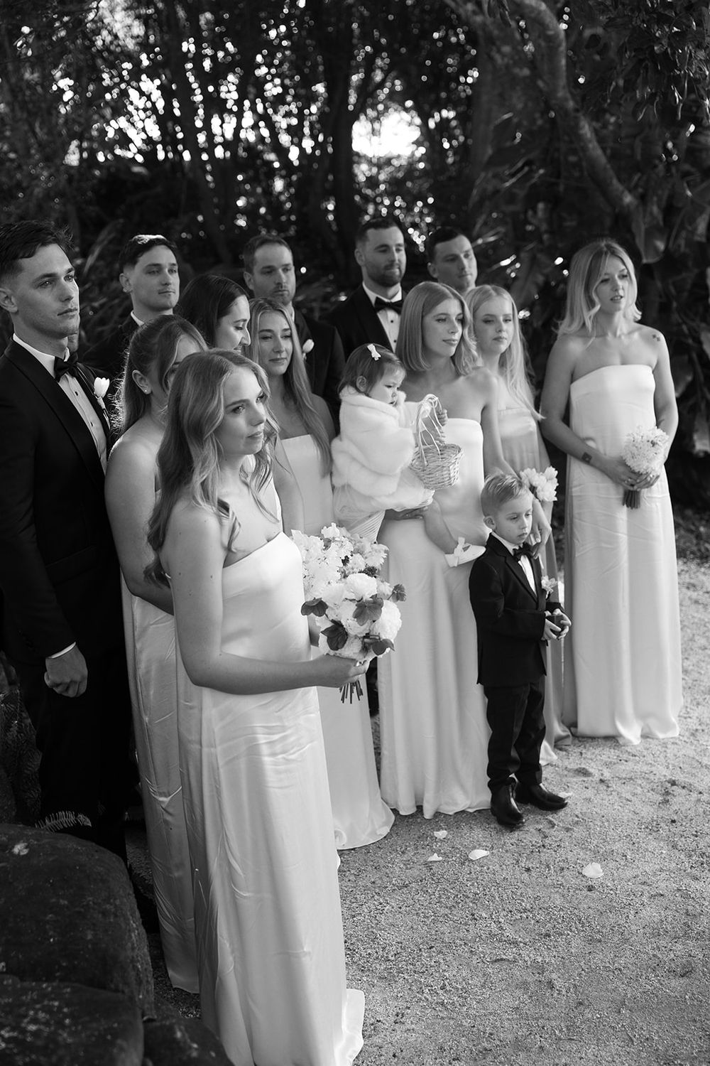 Black and white wedding party photo with bridesmaids in white gowns, groomsmen in tuxedos, and a young ring bearer outdoors.