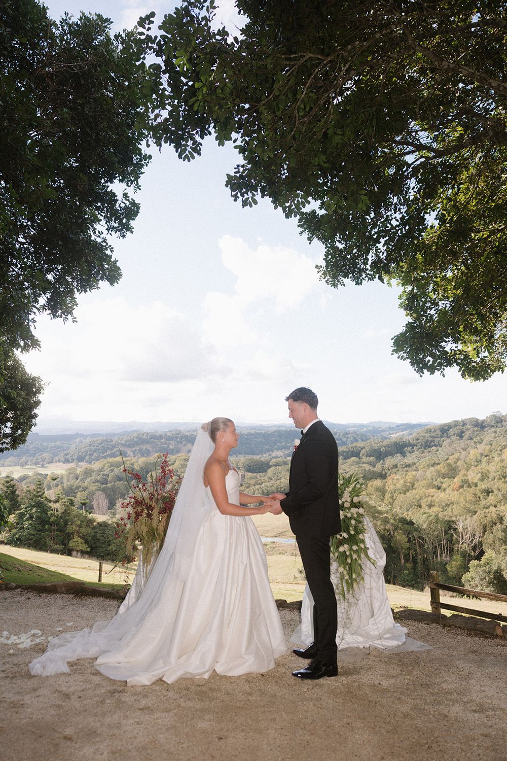 Bride and groom exchange vows under a large tree with rolling green hills in the background on their wedding day.