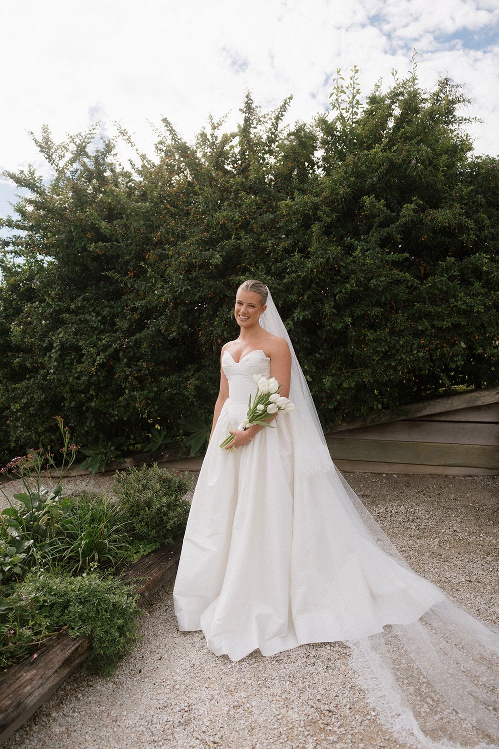 Bride in strapless white ball gown with veil holds white tulip bouquet, posing on gravel path with lush green hedges.