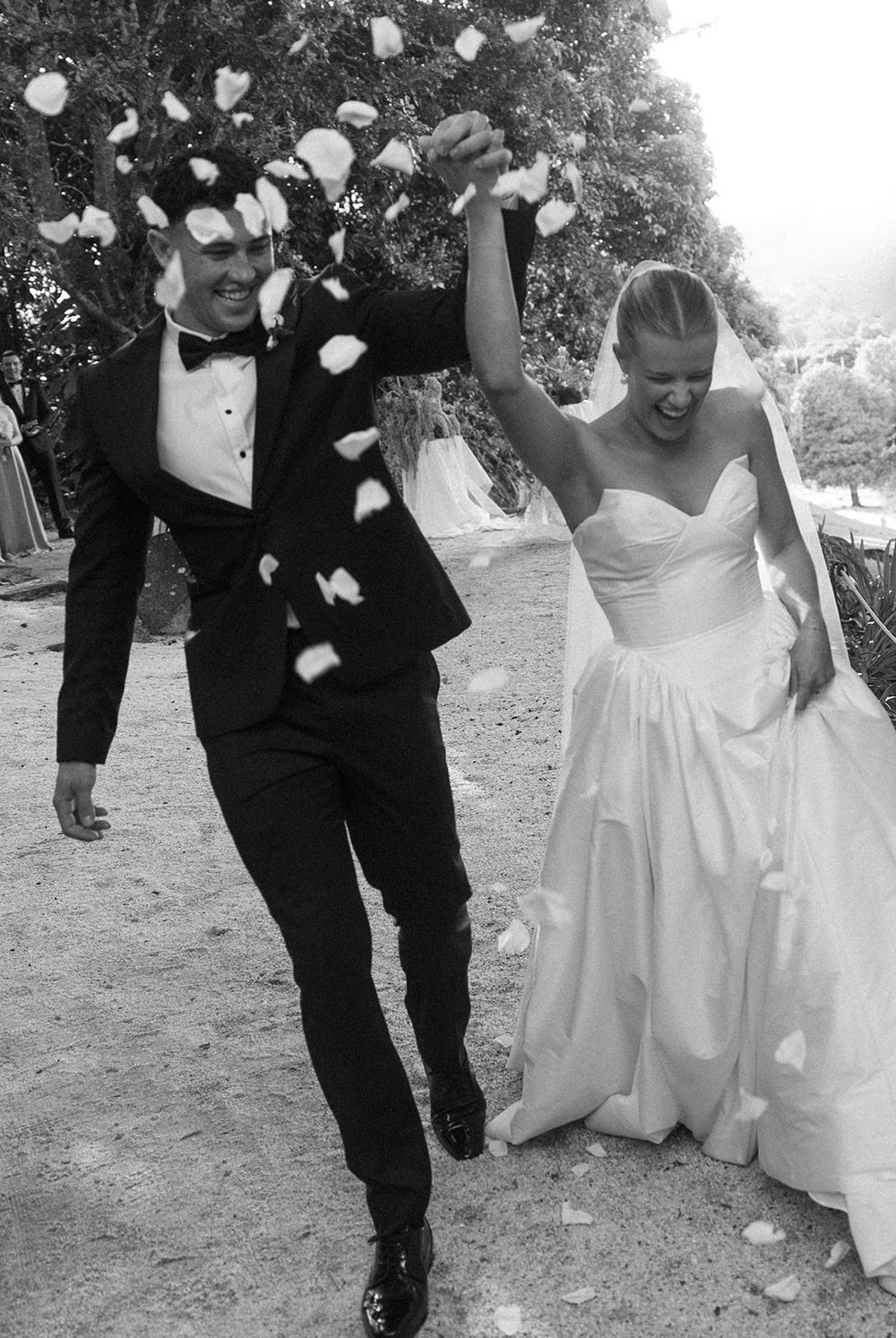 Black and white photo of a joyful bride and groom walking through a shower of rose petals at their wedding.