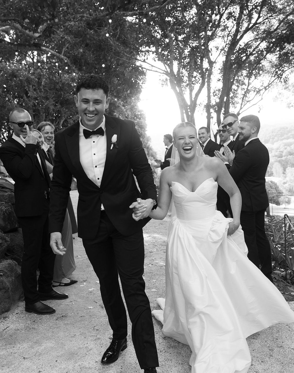 Black and white photo of a joyful couple in wedding attire walking together, surrounded by smiling guests outdoors.