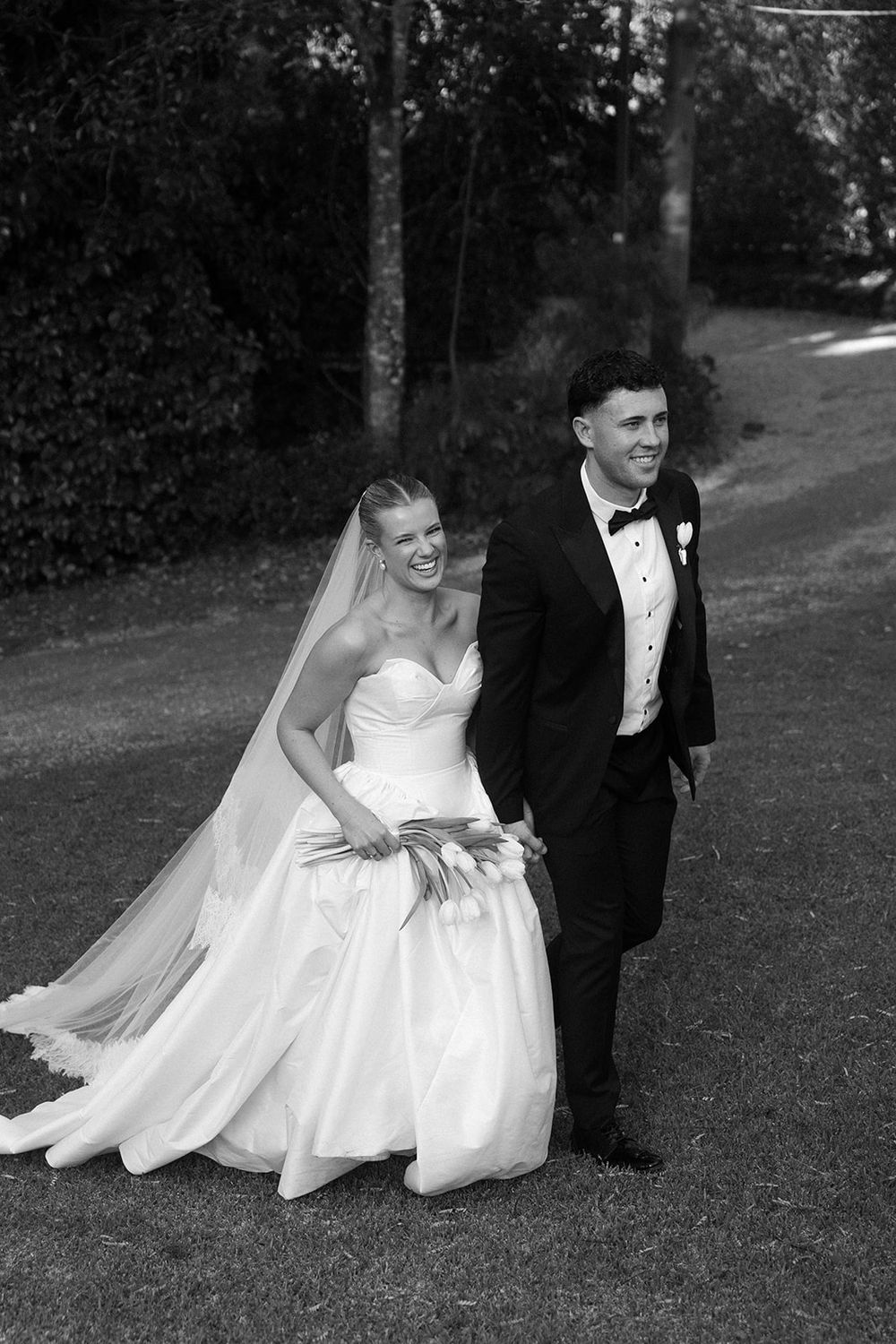 Black and white photo of a smiling bride in a strapless gown and groom in a tuxedo walking together outdoors.