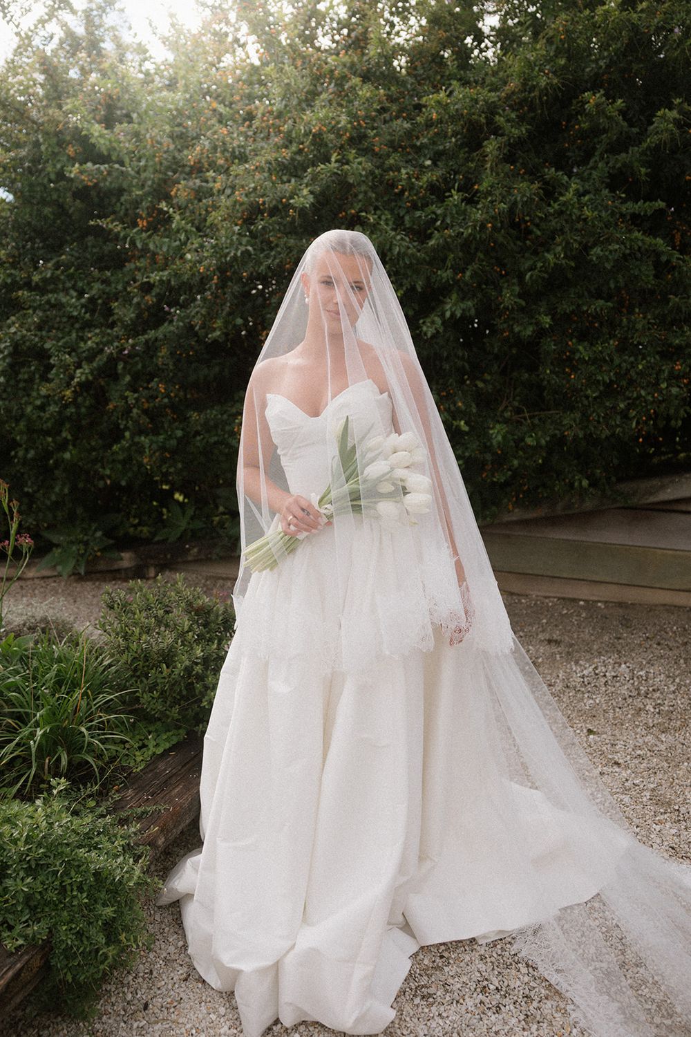 Bride in strapless white tiered gown with long cathedral veil holding white bouquet in lush garden setting.
