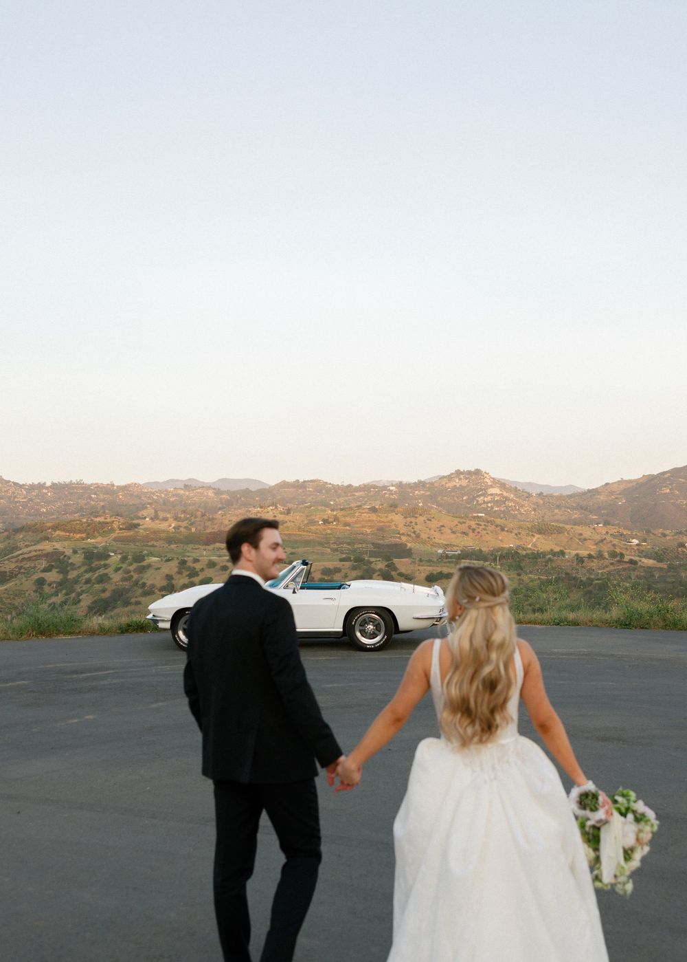 Bride and groom holding hands walking toward a white vintage convertible with scenic hills at sunset.