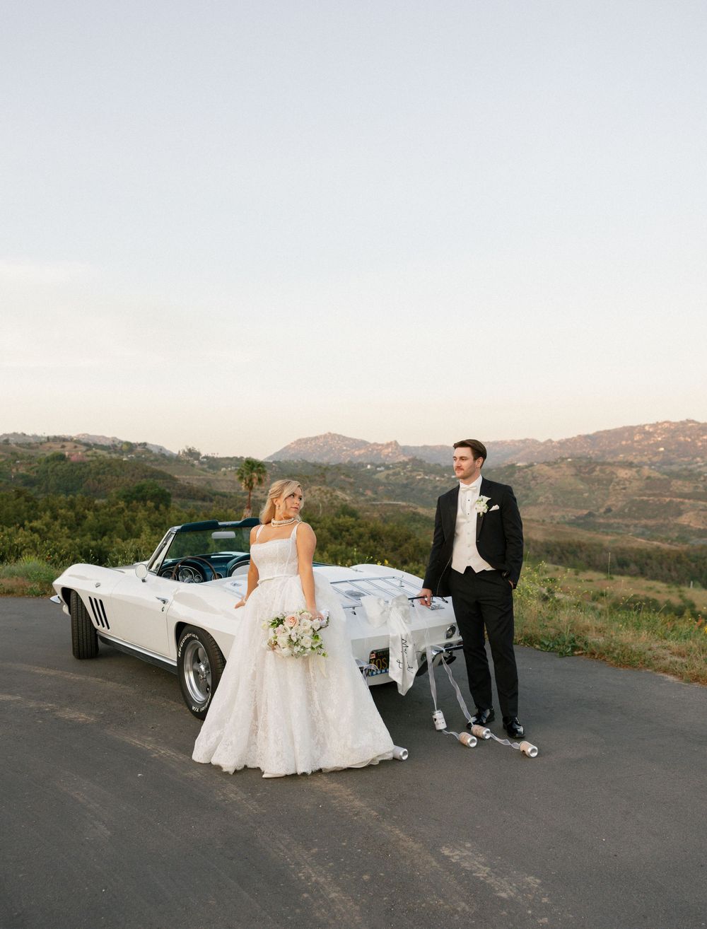 Bride and groom posing beside a classic white Corvette convertible with rolling hills at dusk.