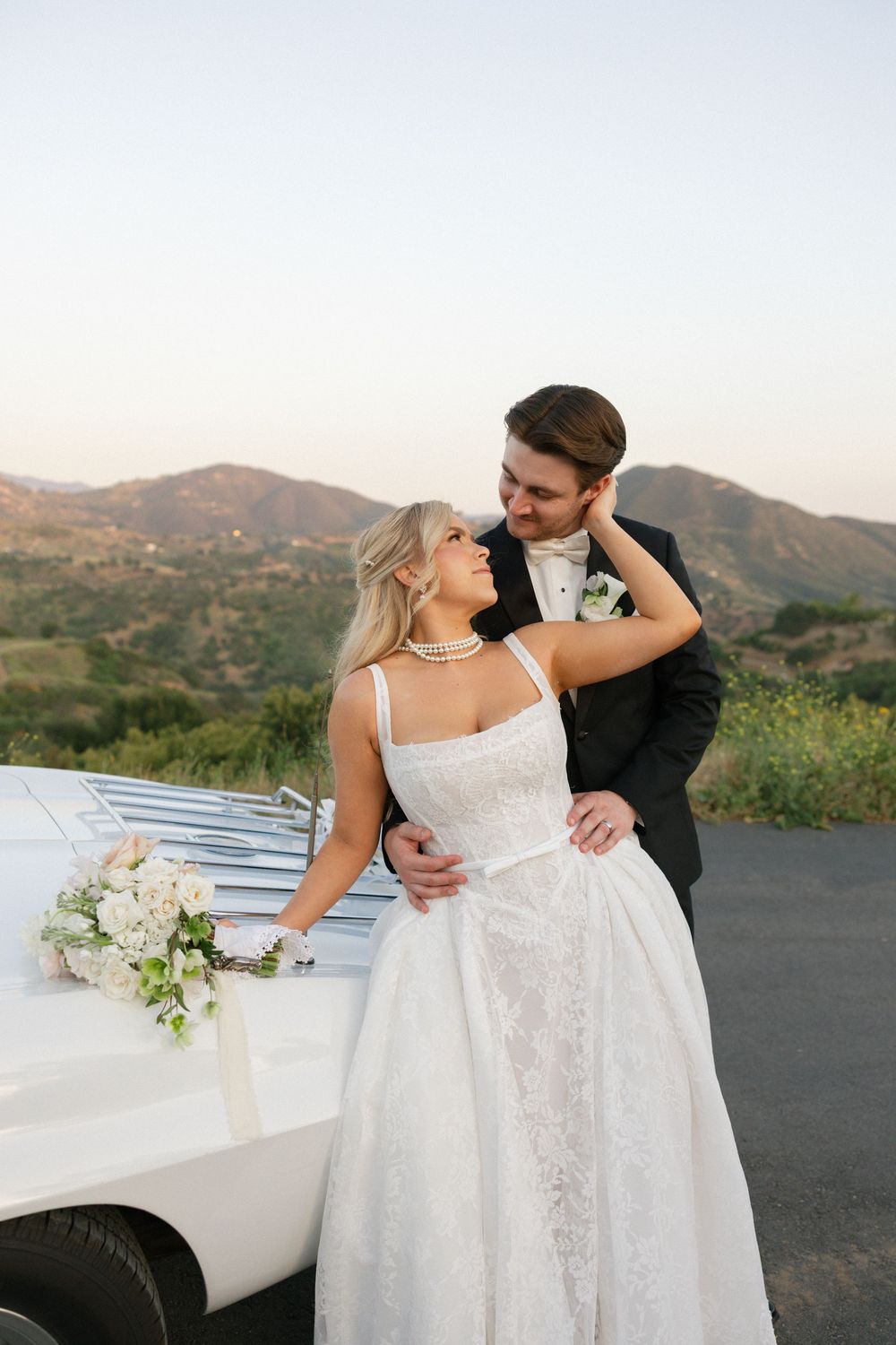 Bride and groom embrace near a white car with mountain views at golden hour, bride holding white floral bouquet.