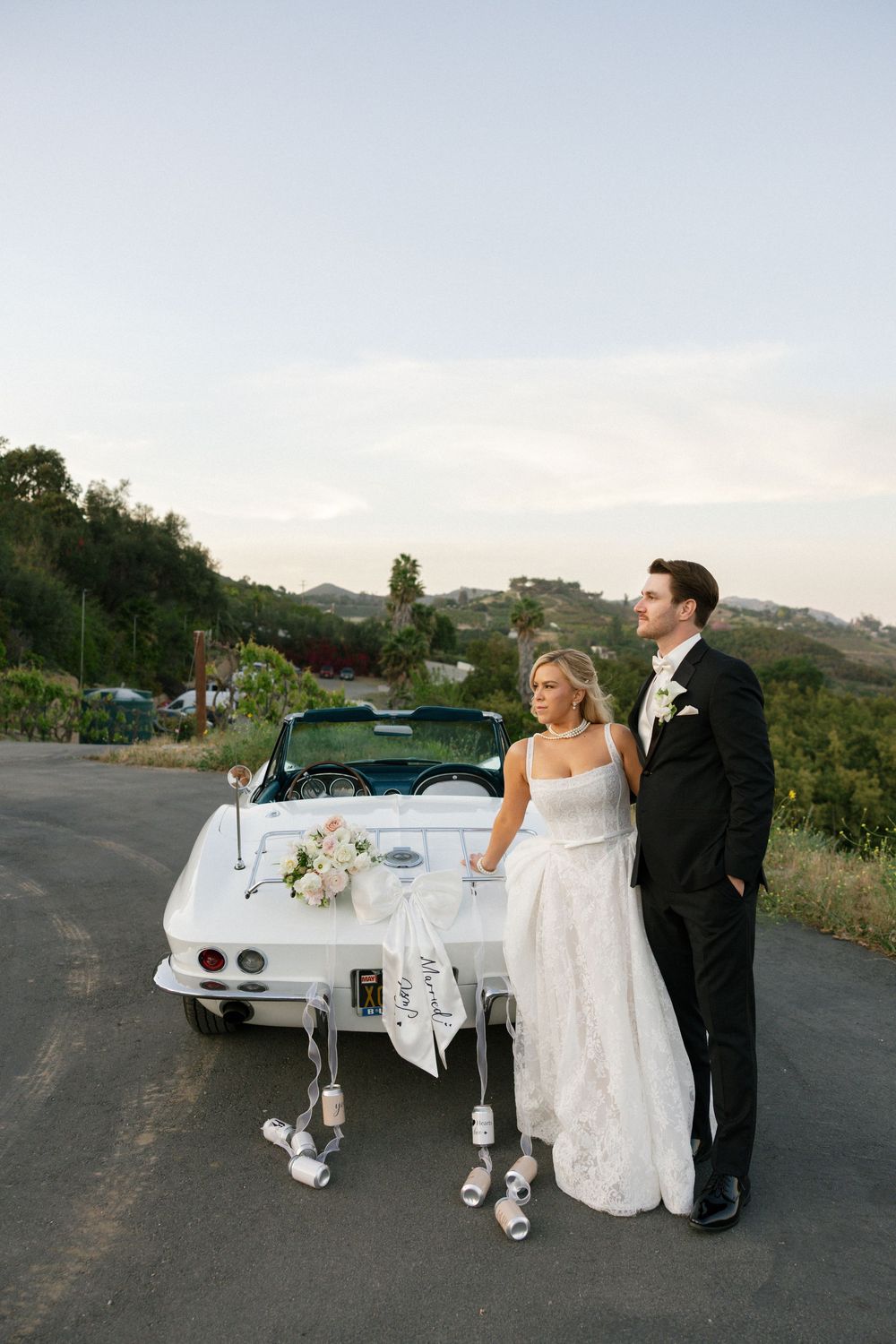 Bride and groom pose beside vintage white Corvette convertible with floral decorations at sunset wedding.