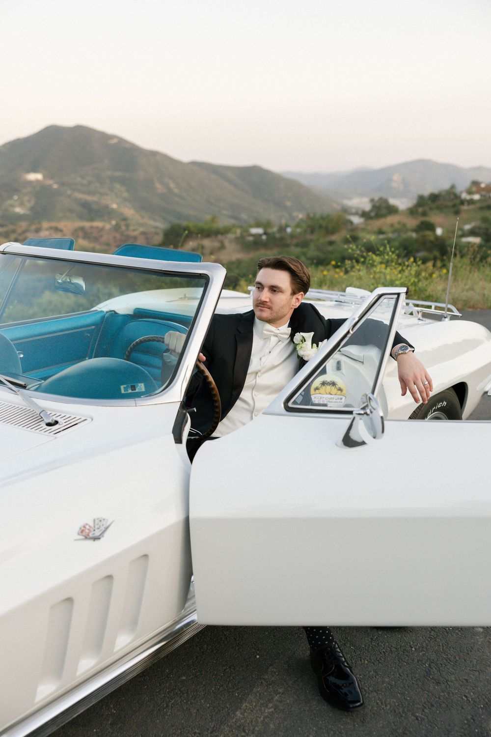 Groom in black tuxedo leaning against white vintage convertible Corvette with mountain backdrop at sunset.