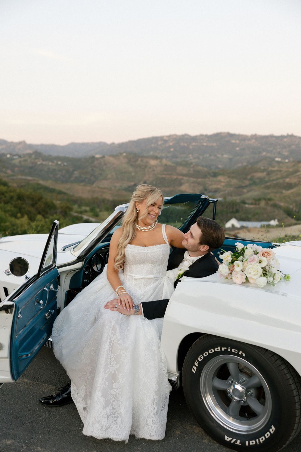 Bride and groom share a romantic moment beside a classic white convertible with rolling hills at sunset.