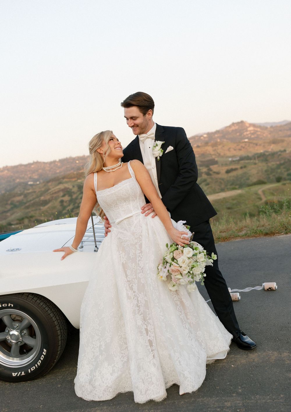 Bride in lace gown and groom in black tuxedo pose together leaning on a white classic car at golden hour.