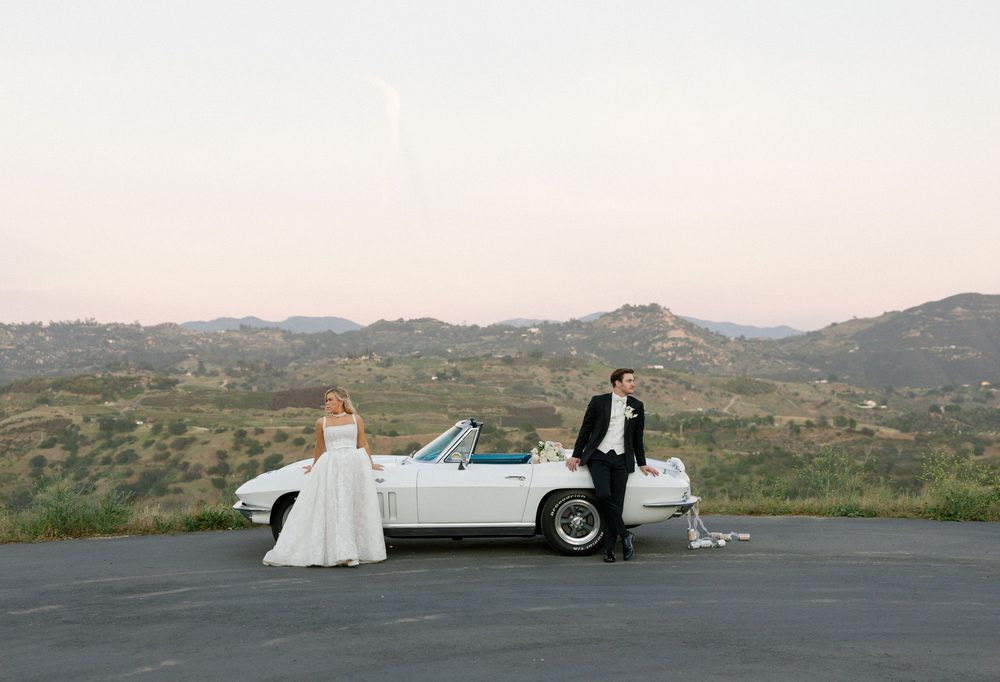 Bride and groom posing beside a classic white convertible Corvette with scenic hills at sunset.