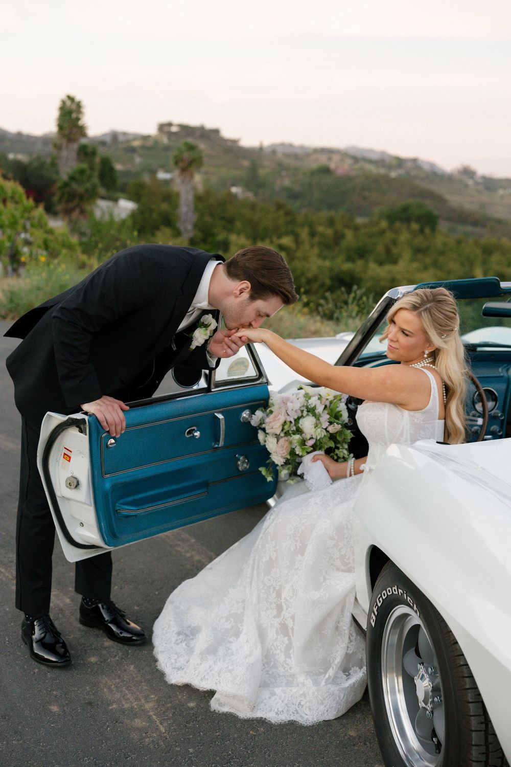 Groom kisses bride's hand as she sits in a vintage white Corvette convertible at dusk.