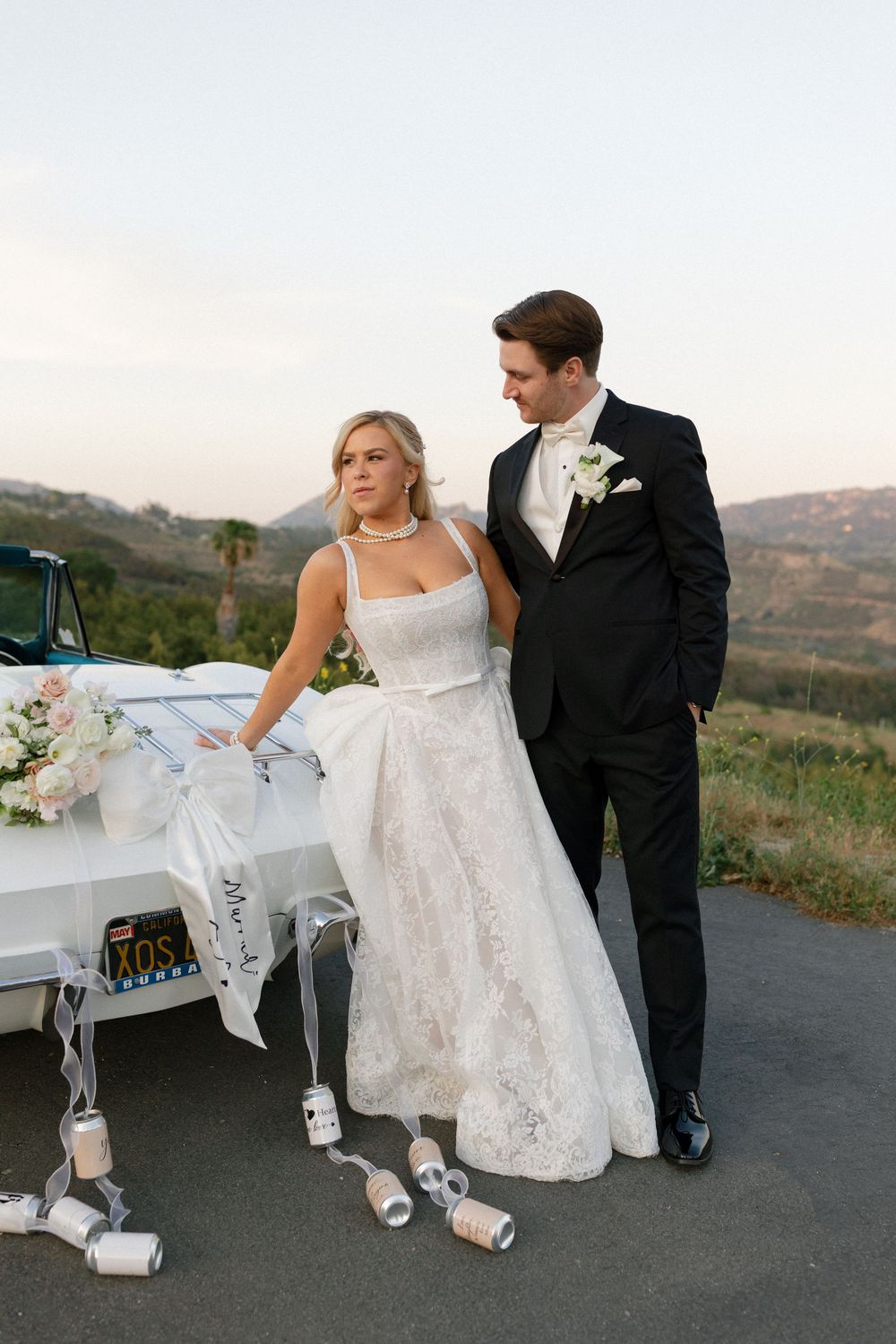Bride in lace gown and groom in black tuxedo pose beside a decorated white wedding car with tin cans at sunset.