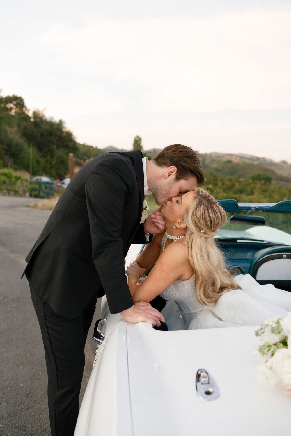 Bride and groom share a romantic kiss beside a white vintage convertible car on their wedding day.