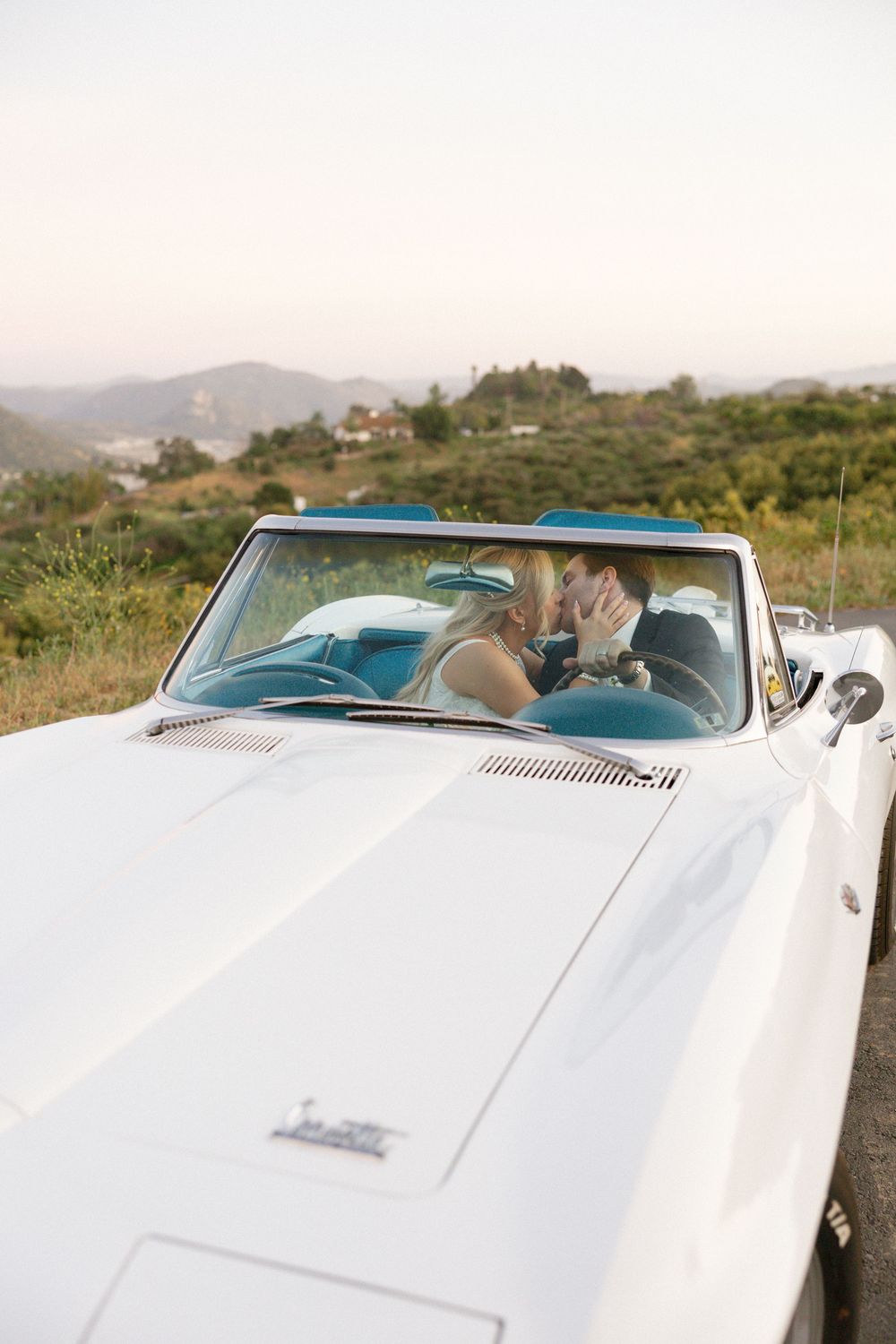 Newlyweds share a kiss inside a classic white convertible with scenic hills in the background.