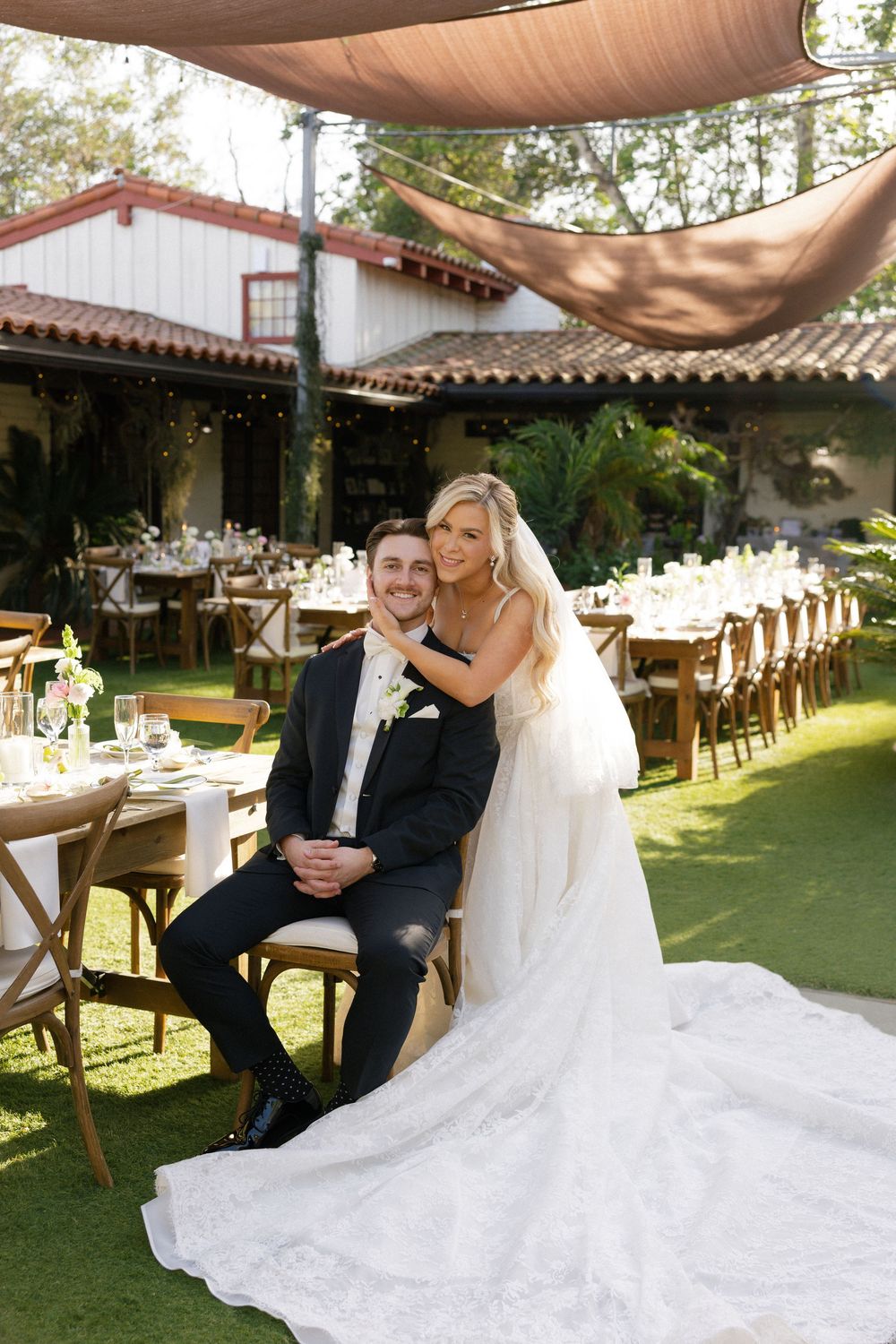 Bride in white lace gown embraces groom in black tuxedo at elegant outdoor wedding reception with string lights.