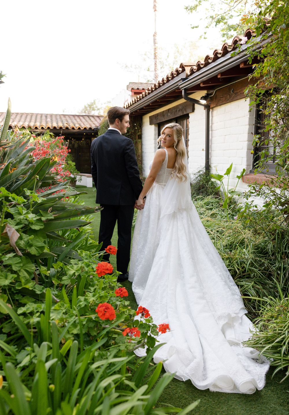 Bride and groom hold hands in a lush garden with red flowers beside a rustic Spanish-style building.
