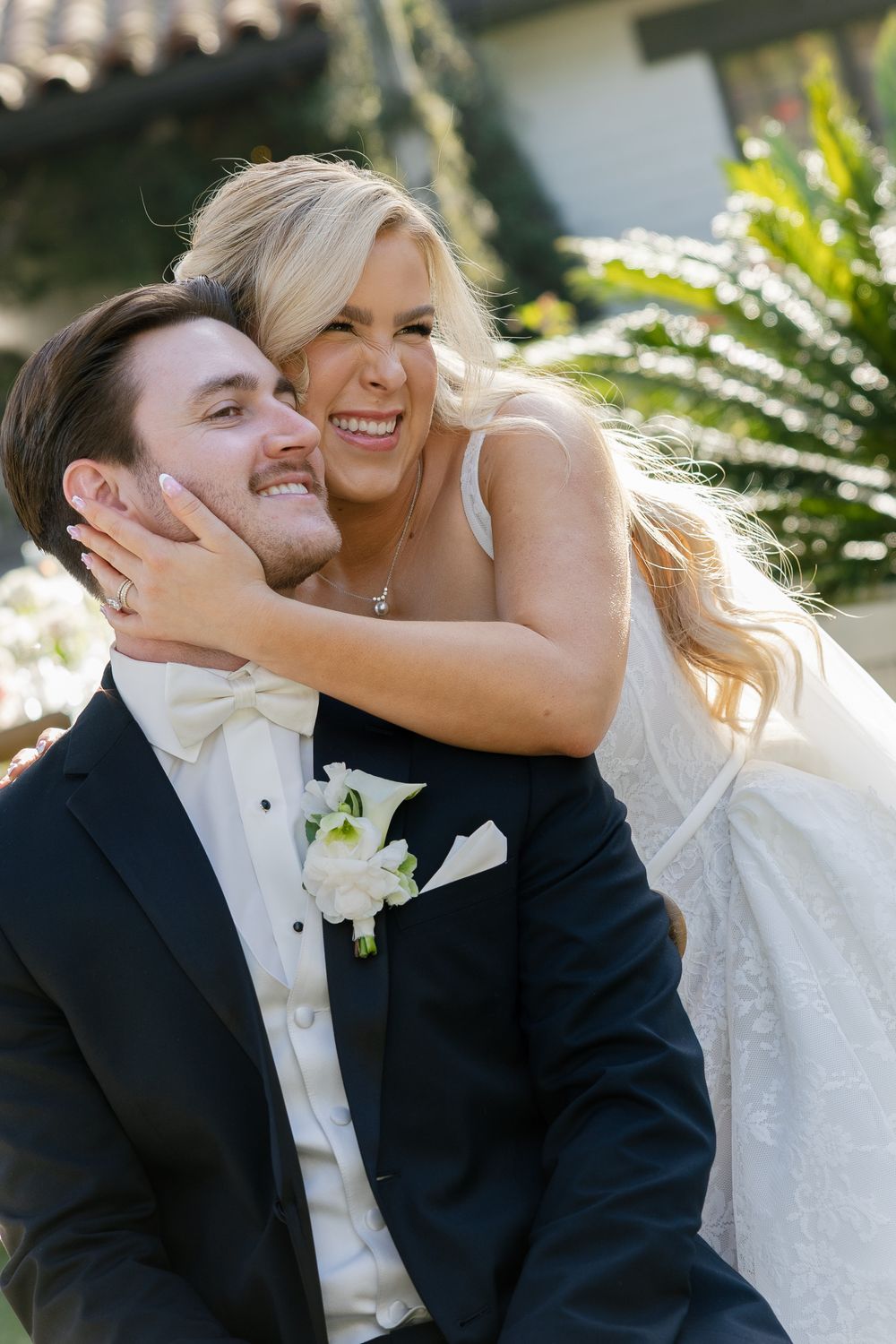 Joyful bride in white gown hugs smiling groom in black tuxedo with white boutonniere outdoors on their wedding day.