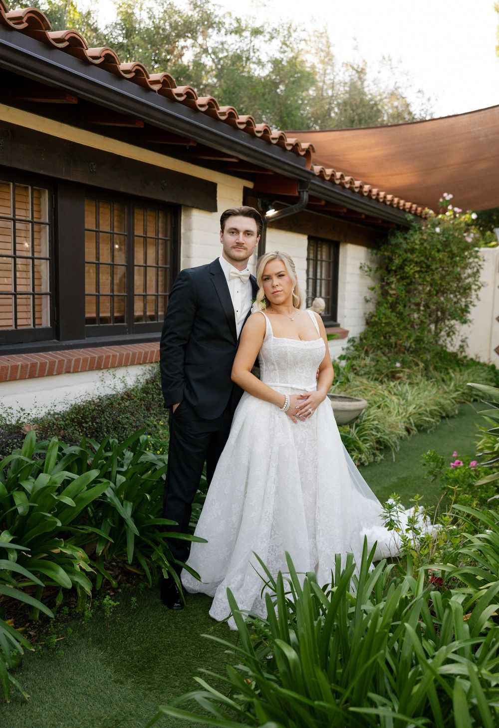 Bride in white ball gown and groom in black tuxedo pose together in lush garden outside Spanish-style venue.