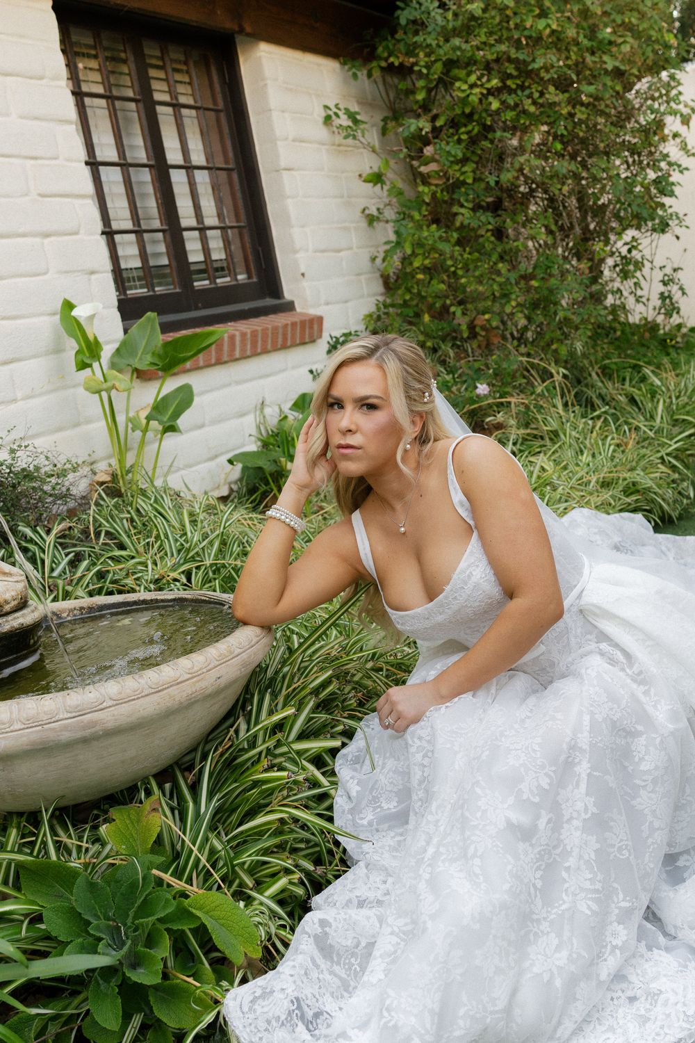 Blonde bride in white lace gown sitting by a stone garden fountain surrounded by lush greenery near a brick building.