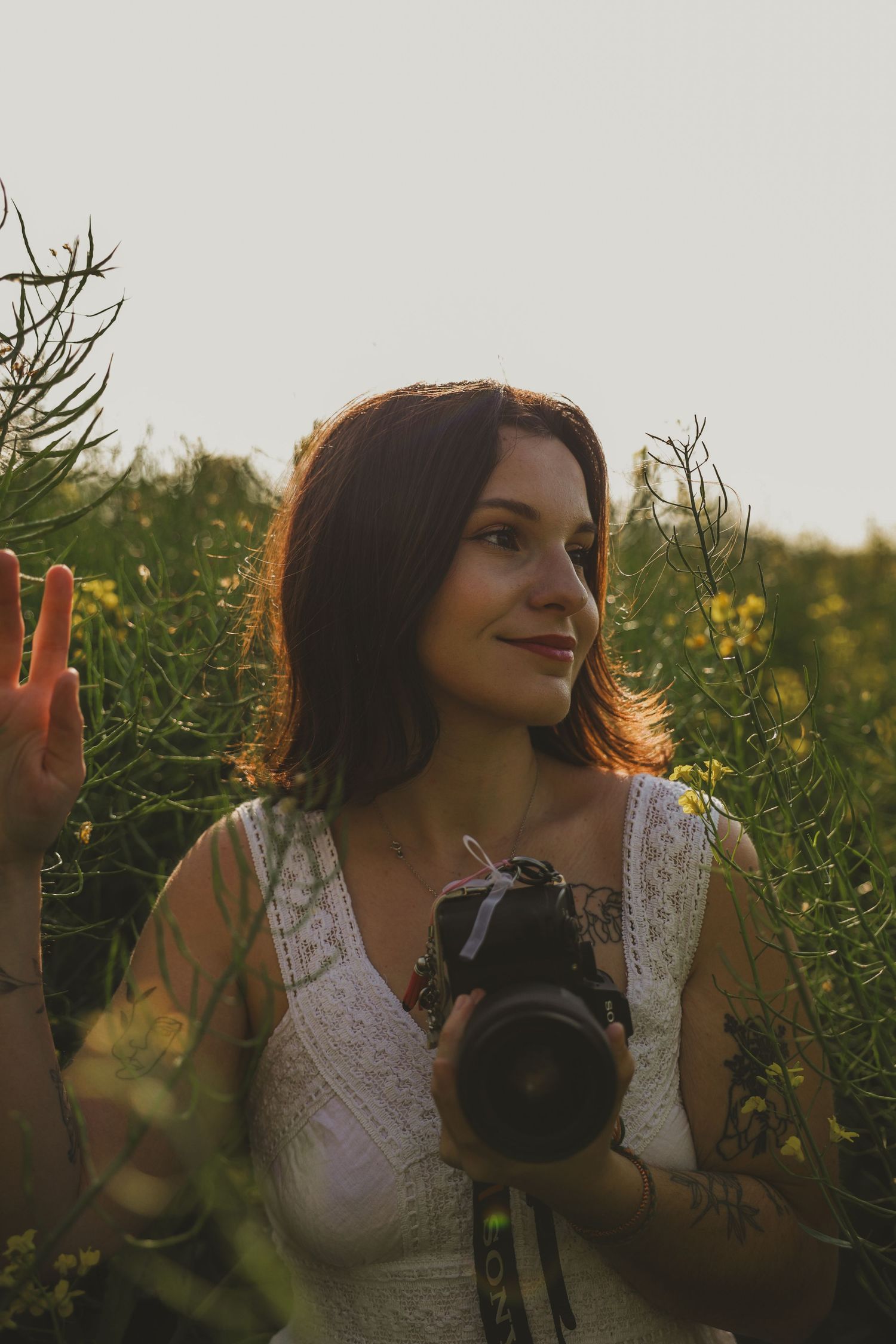 Femme en robe en dentelle blanche tenant une caméra dans un champ ensoleillé de fleurs sauvages au coucher du soleil.