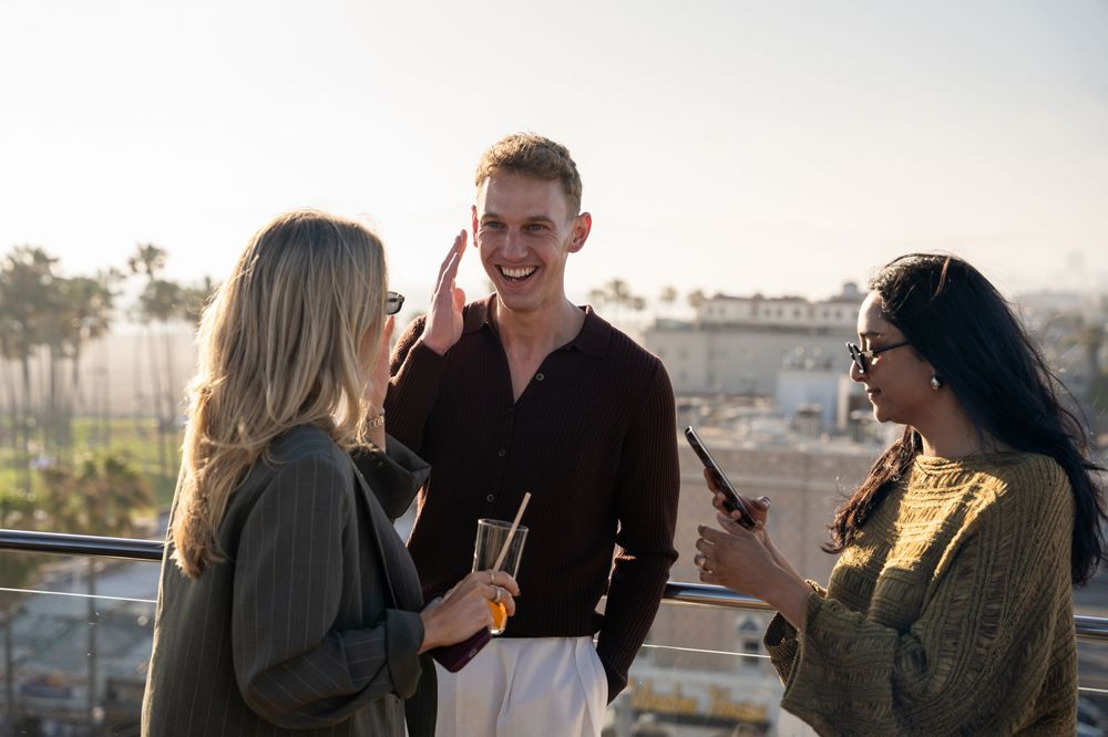 Three young adults socializing on a rooftop terrace at sunset, holding drinks and smiling.