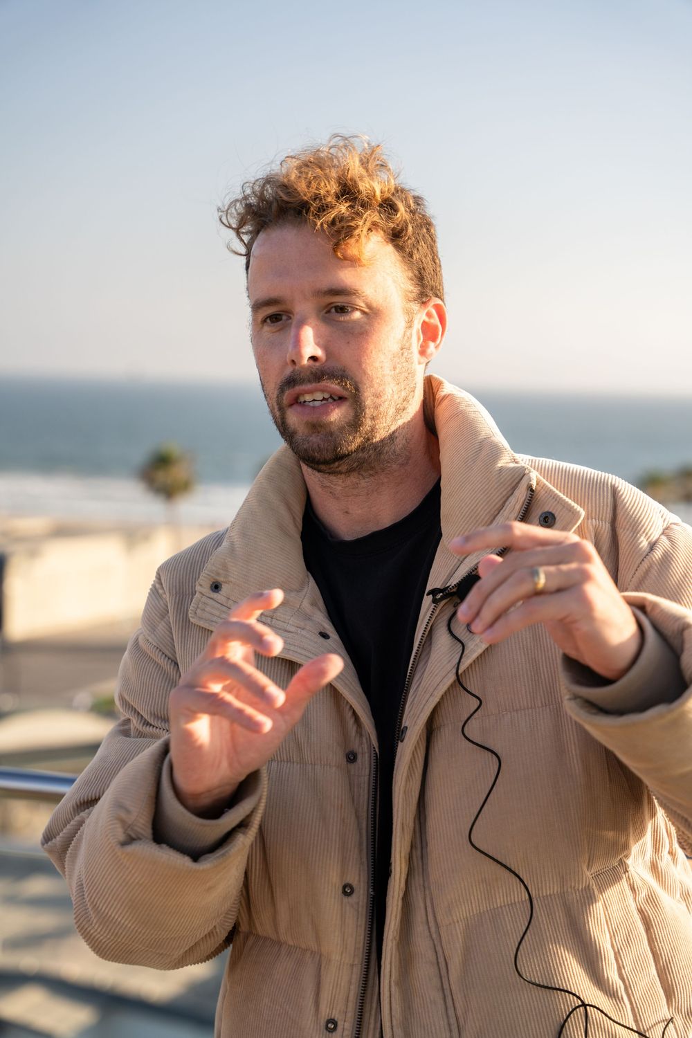 Man in beige puffer jacket gesturing while speaking outdoors near ocean at sunset with lavalier microphone.
