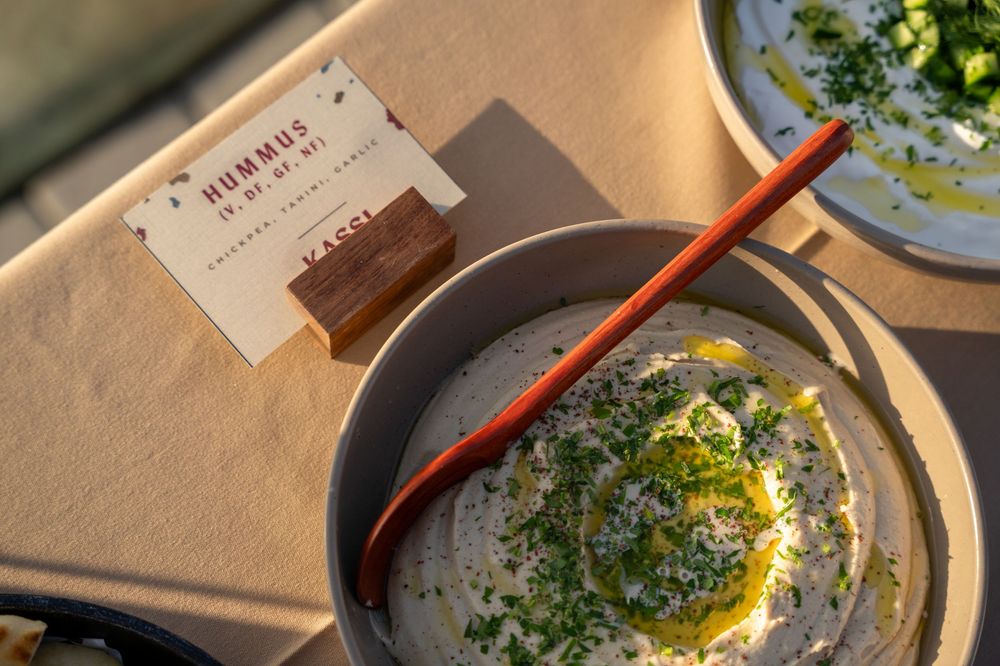 Bowl of creamy hummus topped with olive oil, herbs, and sesame seeds, served with a wooden spoon at Kassl.