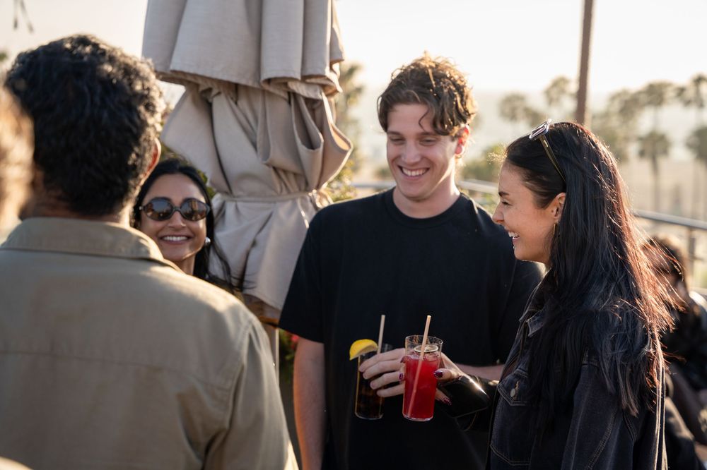 Friends socializing outdoors at sunset, holding colorful cocktails and smiling at a rooftop gathering.