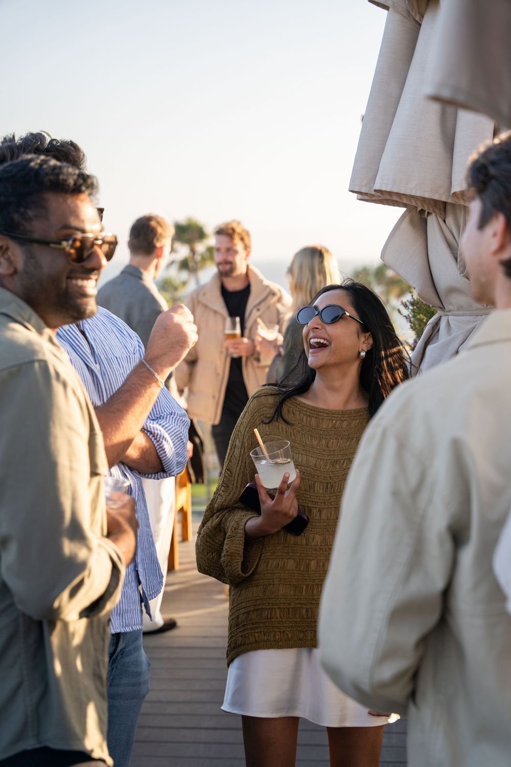 Friends laughing and socializing at an outdoor rooftop party, holding drinks in warm golden sunlight.