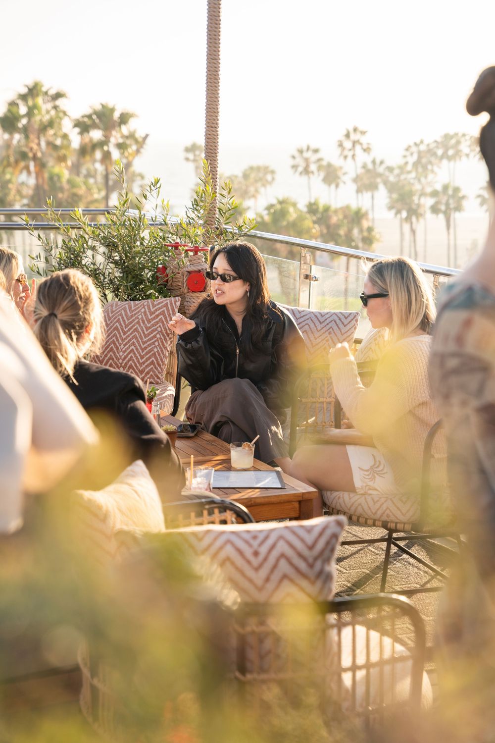 Women socializing on a sunny rooftop terrace with palm trees, enjoying drinks on cushioned chairs in warm golden light.