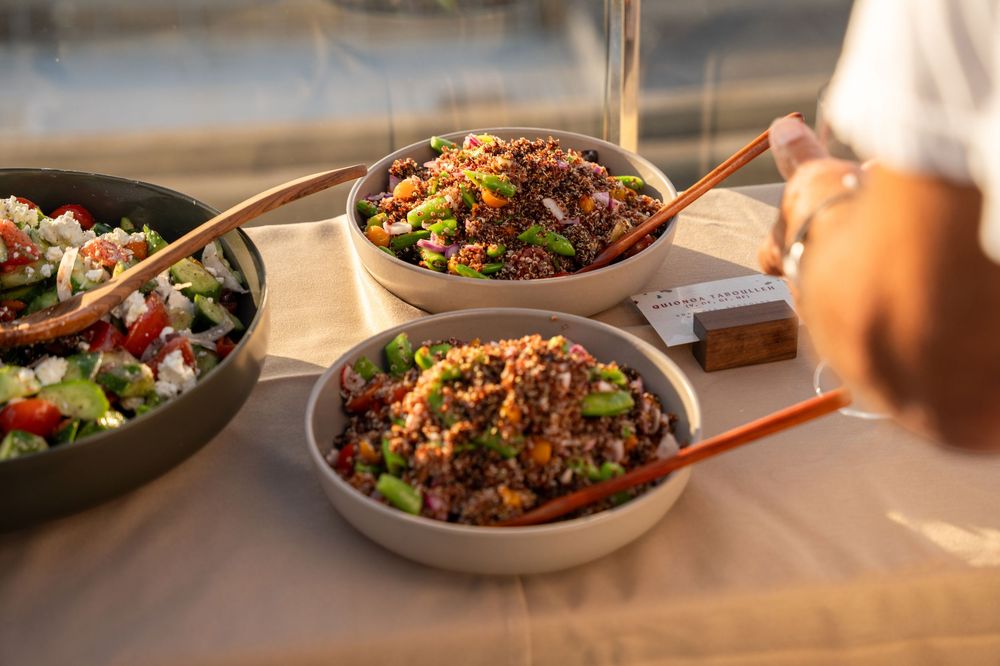 Colorful grain salad bowls with fresh vegetables and wooden serving spoons displayed on a buffet table outdoors.