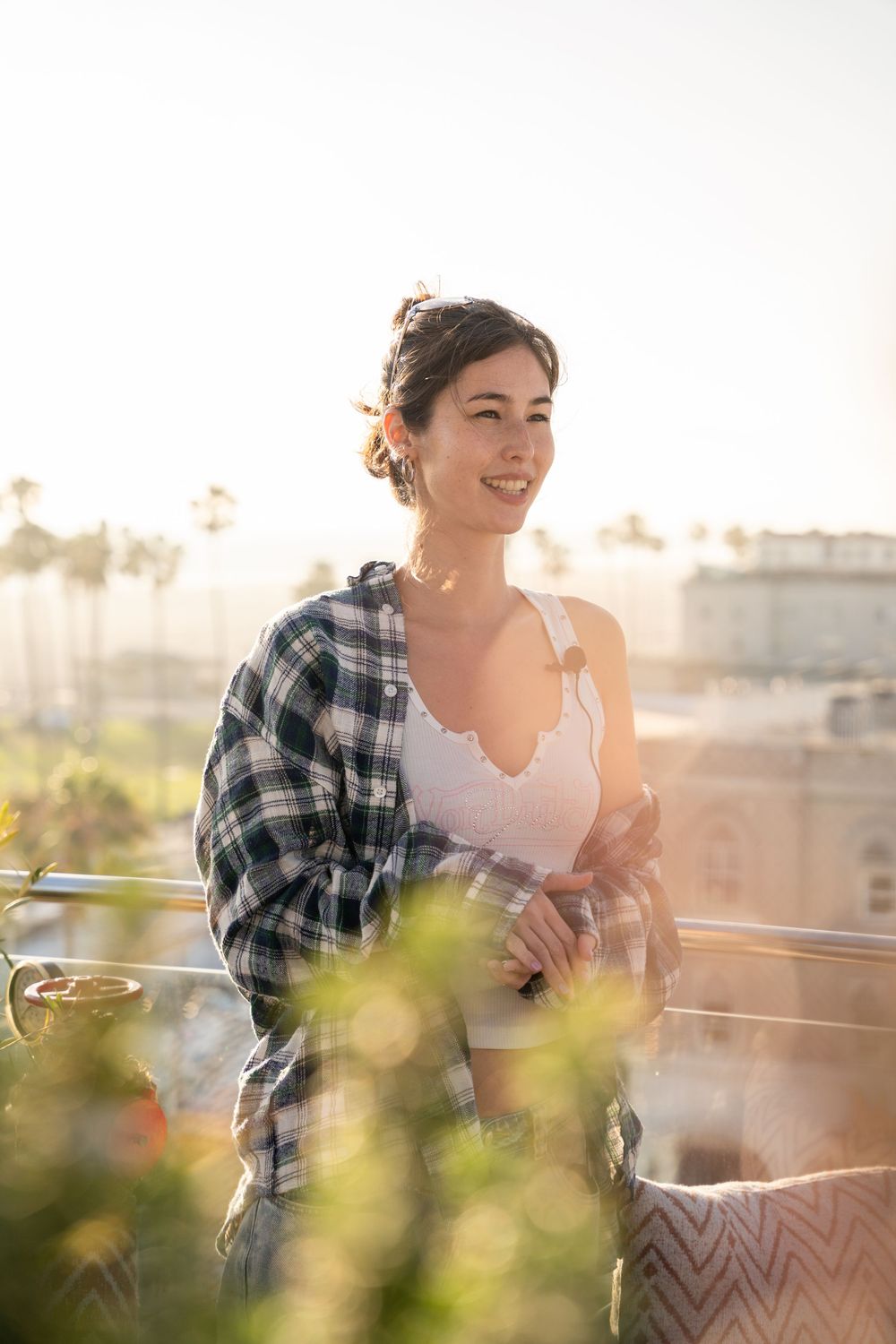 Smiling young woman in plaid shirt leaning on rooftop railing at golden hour with palm trees in background.