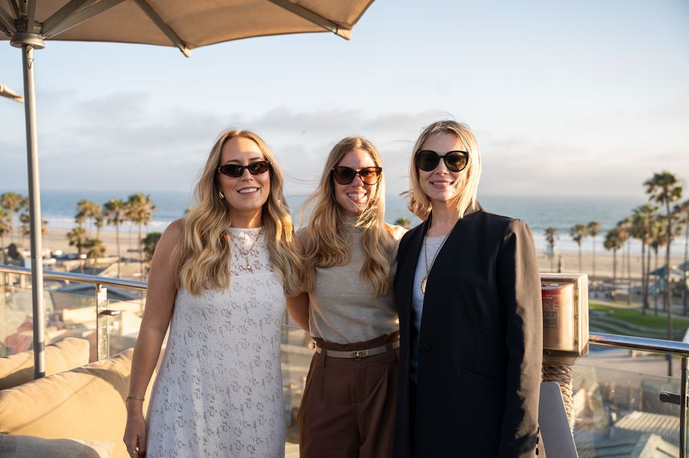 Three smiling women wearing sunglasses pose together on a rooftop terrace with a beach view at sunset.