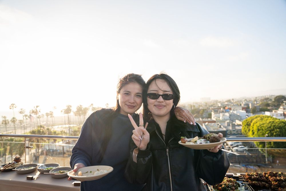Two women posing on a rooftop terrace with plates of food, flashing peace signs with a coastal city view behind them.