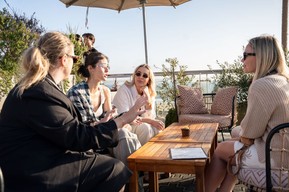 Four women enjoy sunny outdoor patio conversation at a wooden table under an umbrella with ocean views.