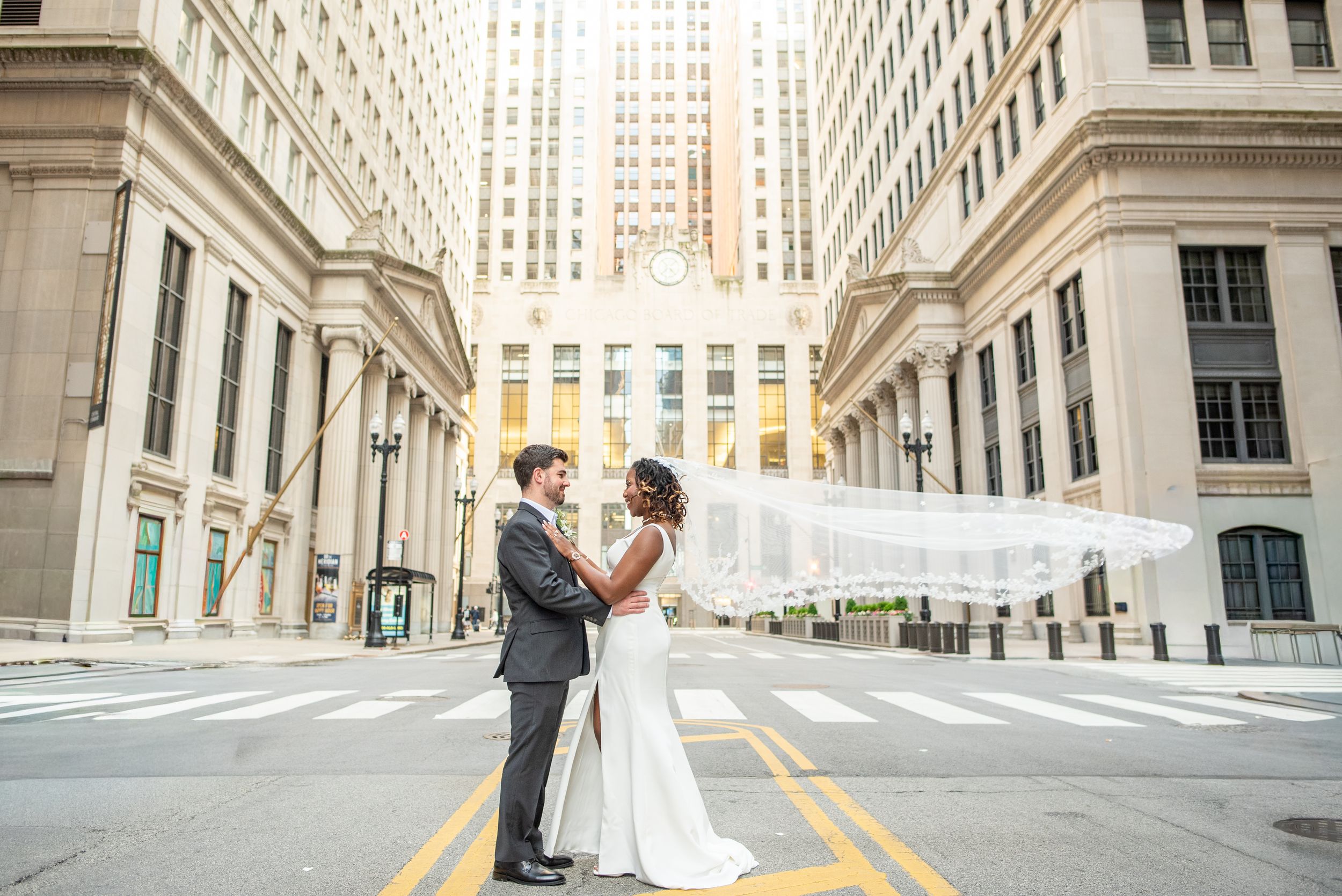 Bride and groom share a romantic moment on a city street, with the bride's veil flowing in the wind.