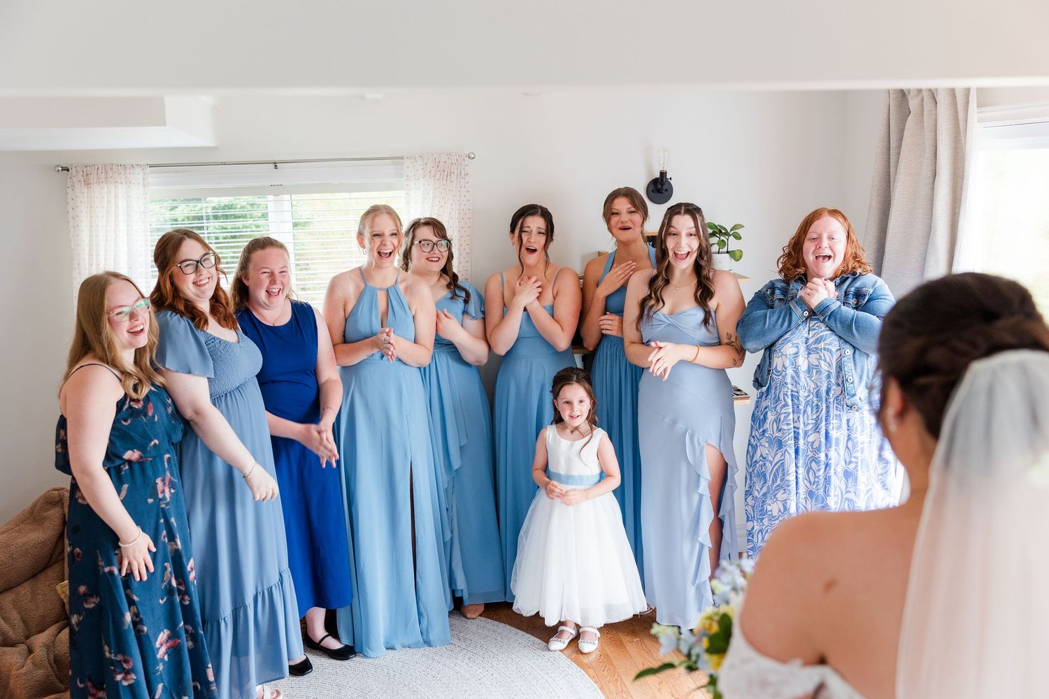 Bridesmaids in blue dresses react with joy and excitement as bride reveals her wedding gown during bridal preparations.