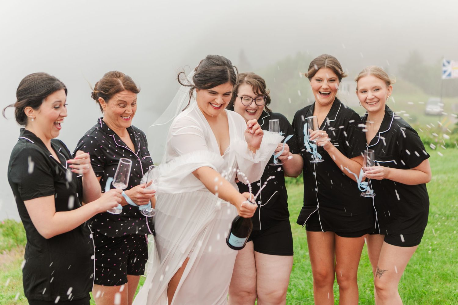 Bride in white robe pops champagne with bridesmaids in black pajamas outdoors on a misty mountain wedding day.