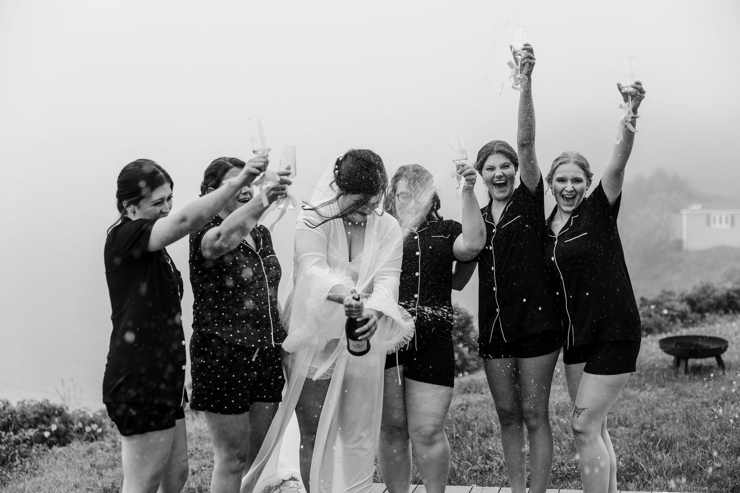 Bride pops champagne with bridesmaids in pajamas celebrating outdoors in black and white wedding photo.