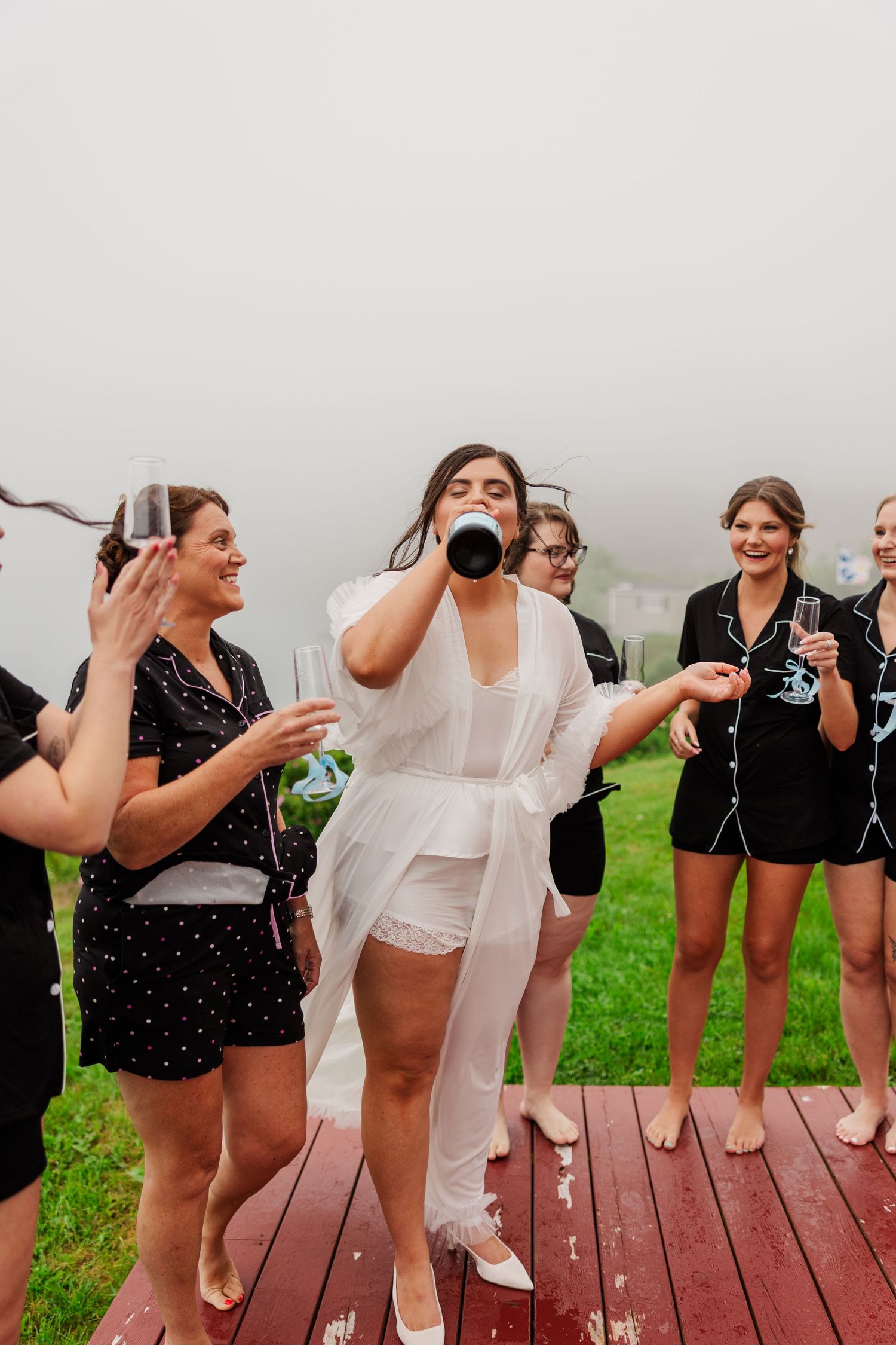 Bride in white robe drinks champagne bottle with bridesmaids in black pajamas on foggy outdoor deck.