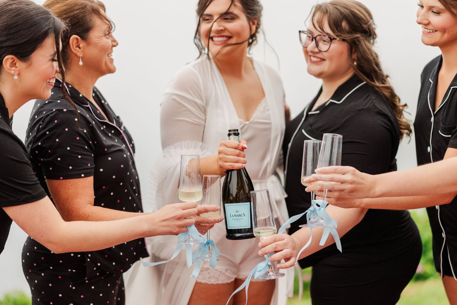 Bride in white robe celebrates with bridesmaids in black, toasting champagne glasses with blue ribbon details.