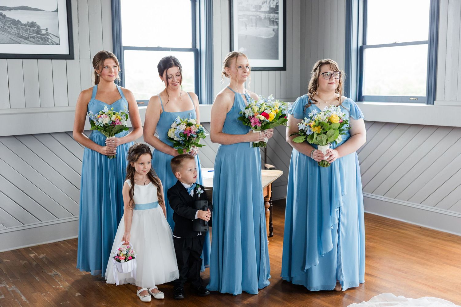 Five bridesmaids in blue gowns hold colorful bouquets alongside a flower girl and ring bearer at a wedding.