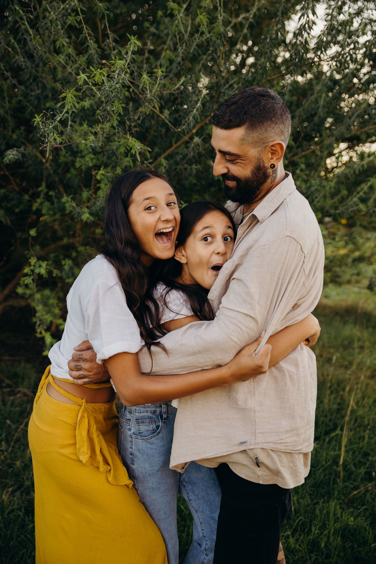 A joyful family of three shares a warm embrace outdoors, laughing together near lush green trees at golden hour.